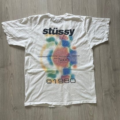 White t-shirt with colorful design and 'stussy' branding on a wooden floor.