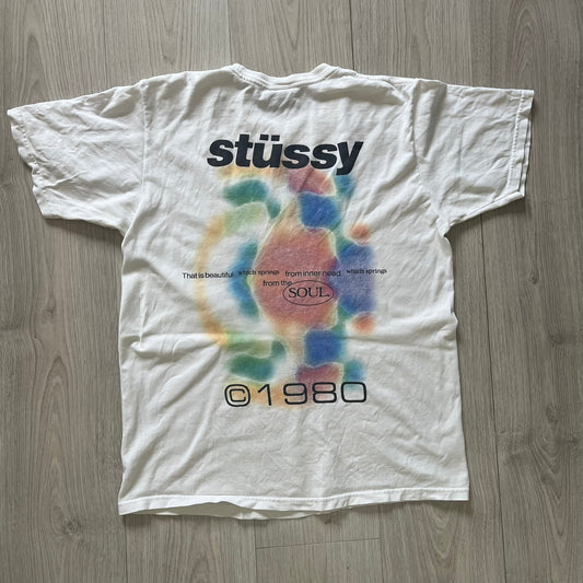 White t-shirt with colorful design and 'stussy' branding on a wooden floor.
