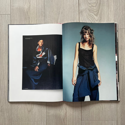 Open book showing two fashion images of a model wearing a black top and blue skirt on a wooden surface.